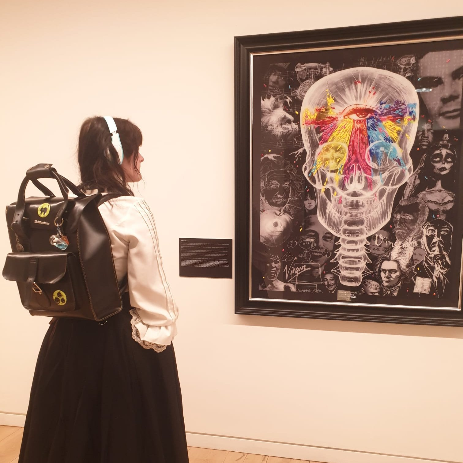 A young woman wearing headphones with a backpack, looking at a framed piece of artwork featuring a colourful skull with black and white faces surrounding it.