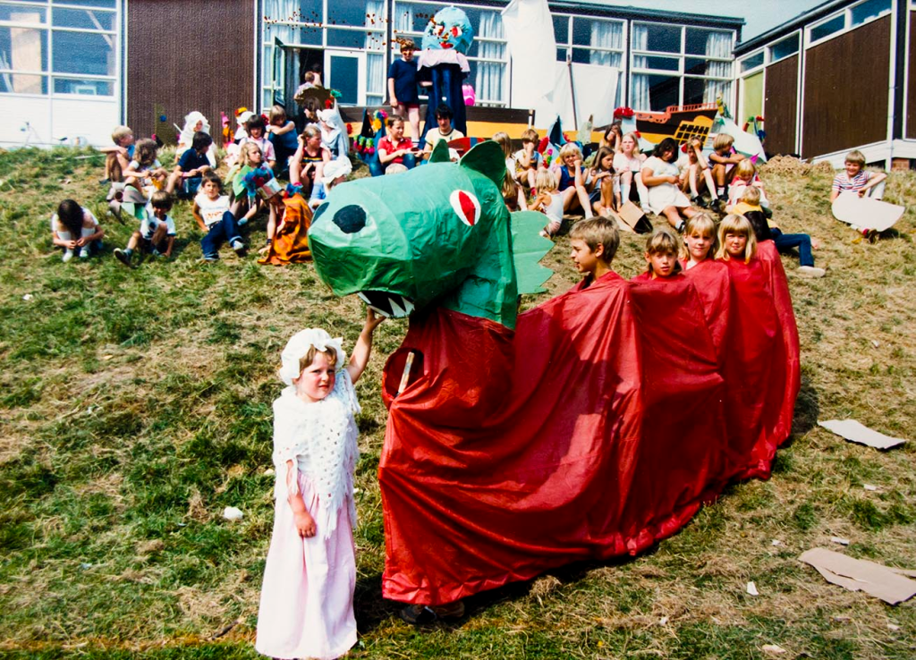 A green and red dragon operated by several children in the body. In the background is a group of children sitting on a hill.