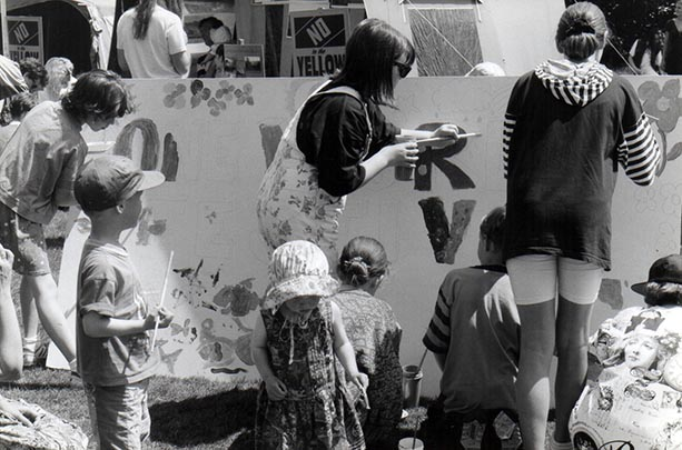 A black-and-white photo of 3 women painting a giant sign; children are by them, also helping. On the board, letters can be seen, but not enough to form a word.