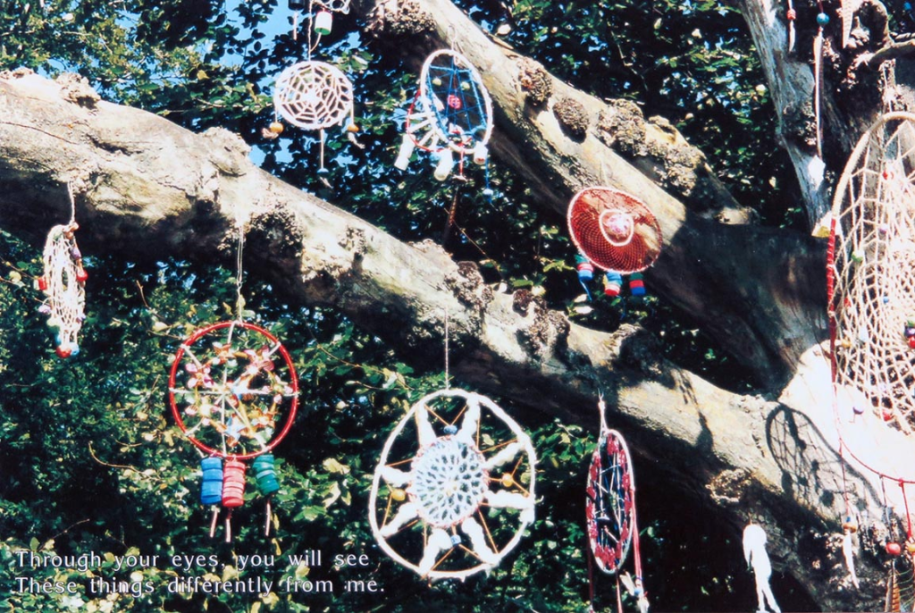 Tree branches decorated with colourful crocheted dream catchers. Text at the bottom states "Through your eyes, you will see. These things differently from me."