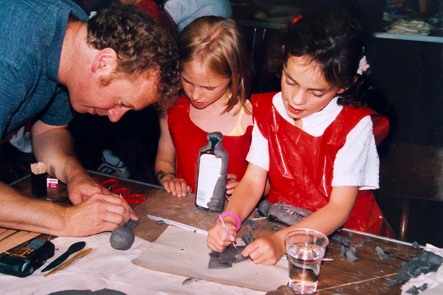2 young girls sat at a table making clay art, a man is leaning over the table helping them.
