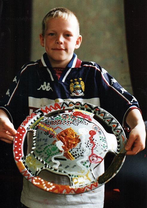 A young boy holding a car wheel trim that's decorated with colours such as red, green, orange and black.