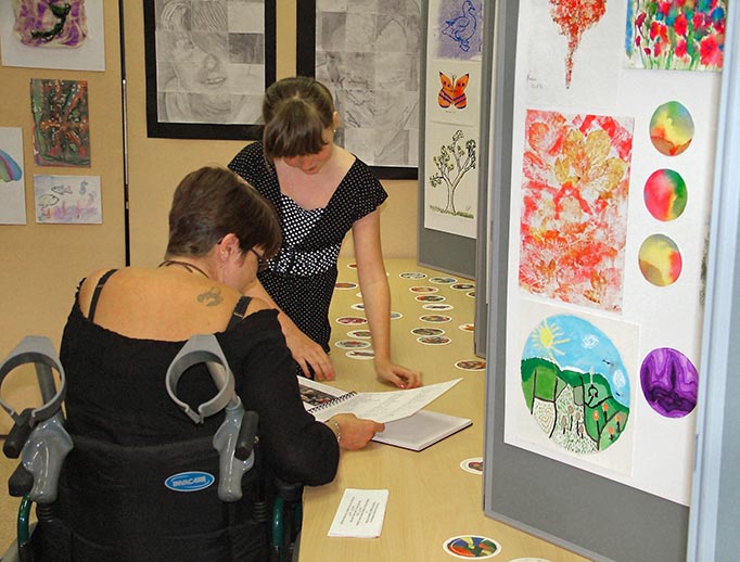 A person in a wheelchair and somebody stood up looking at a book on a table. Around the room is colourful artwork, butterflies, trees and hills.