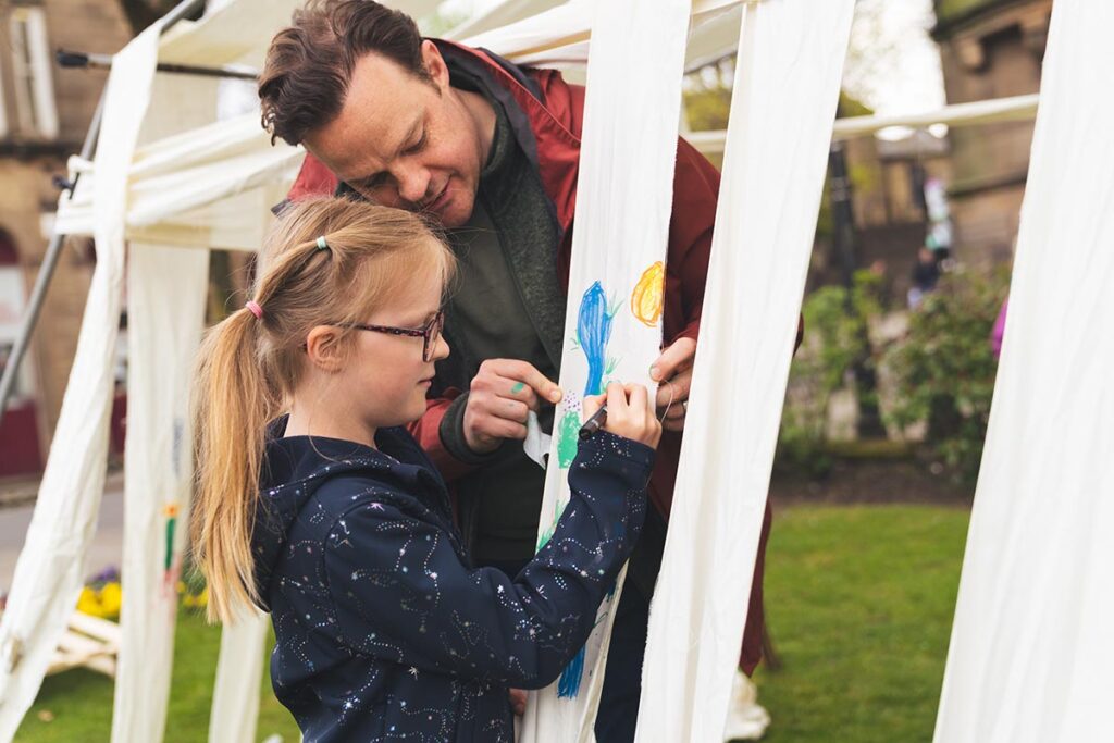 A young girl drawing on a piece of fabric that's part of a tent with a man beside her holding it still.