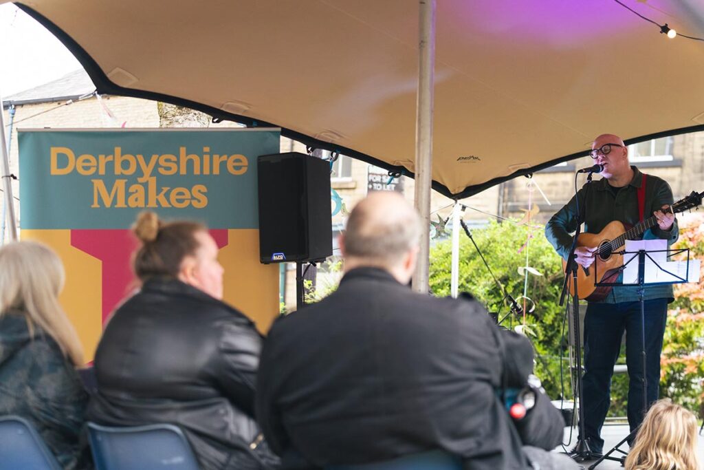 A man stood on a stage playing an acoustic guitar, we see the backs of the audience watching and a sign stating "Derbyshire Makes".