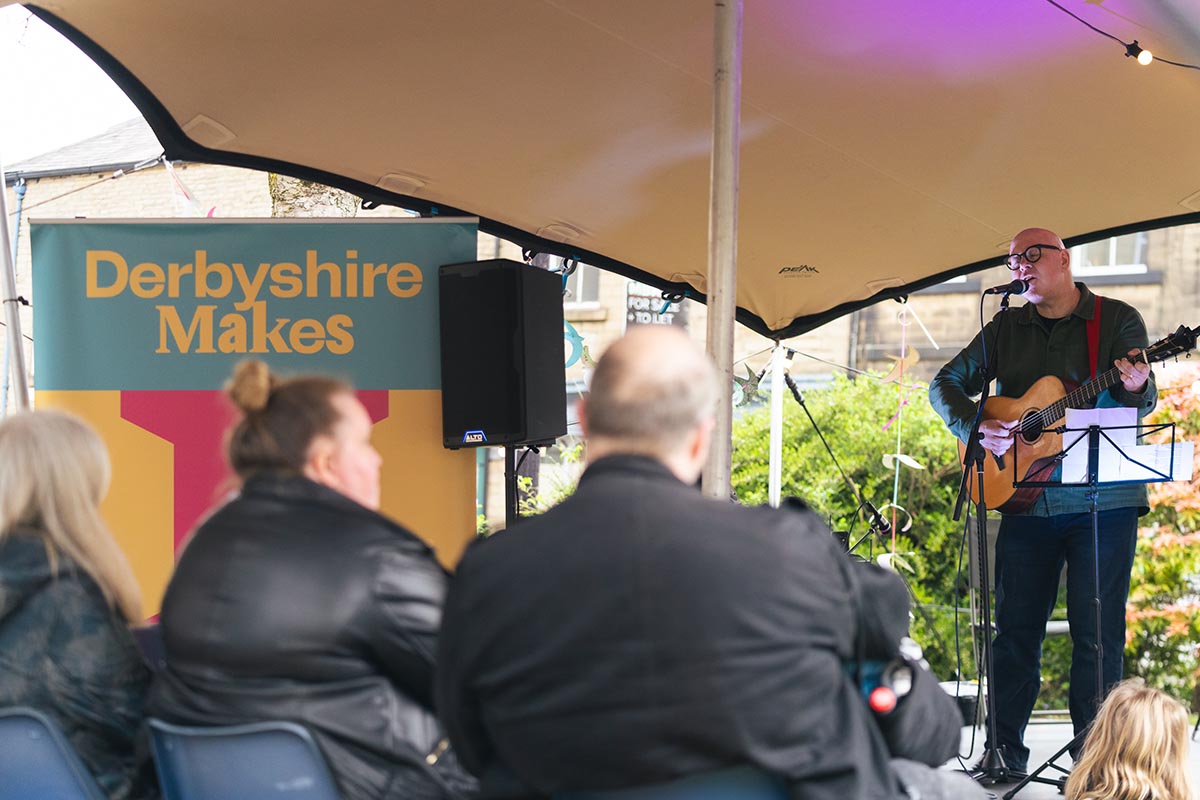 A man stood on a stage playing an acoustic guitar, we see the backs of the audience watching and a sign stating "Derbyshire Makes".