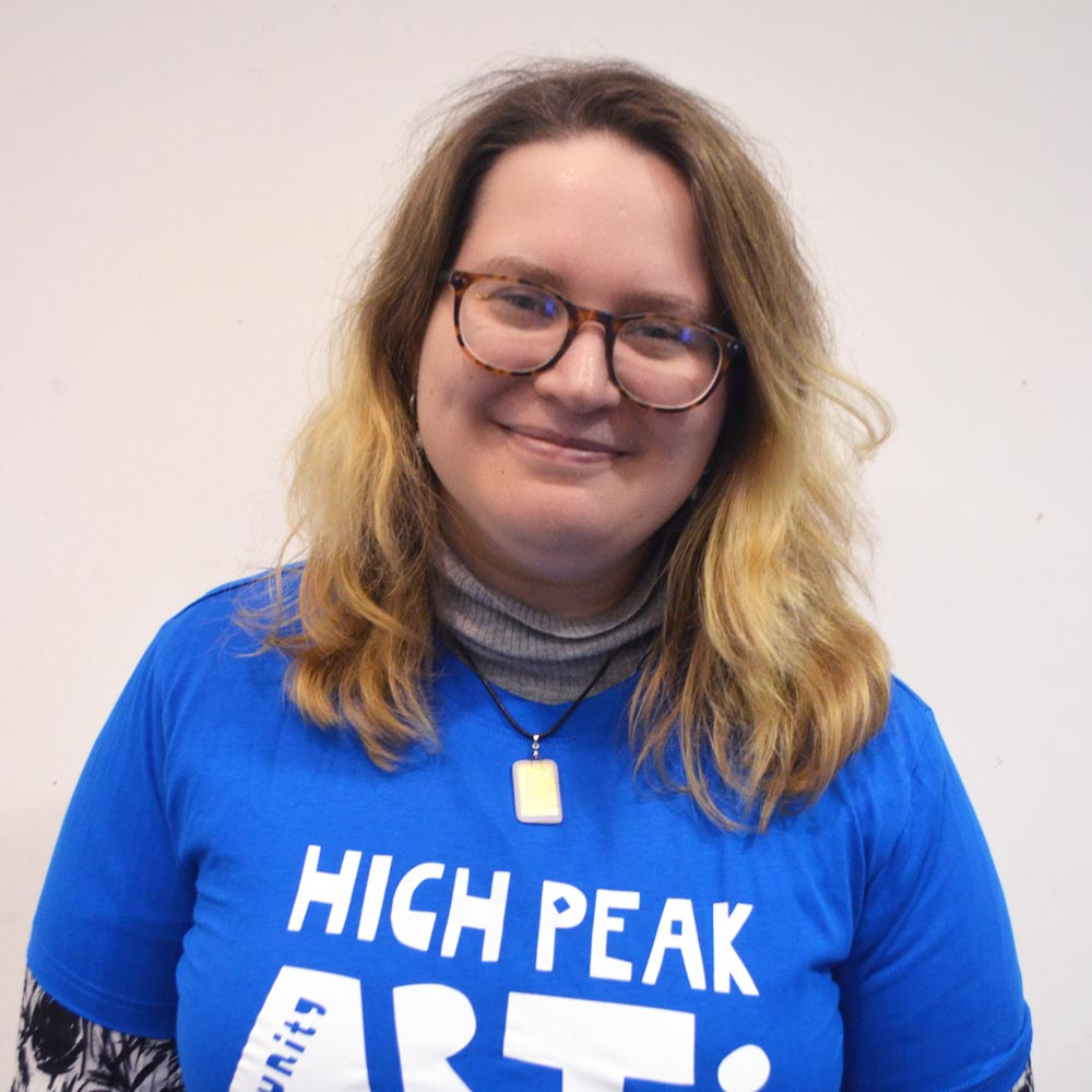 A woman with glasses and short hair wearing a blue High Peak Community Arts logo t-shirt.