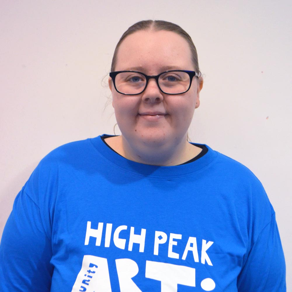 A woman with glasses wearing a blue logo High Peak Community Arts t-shirt.