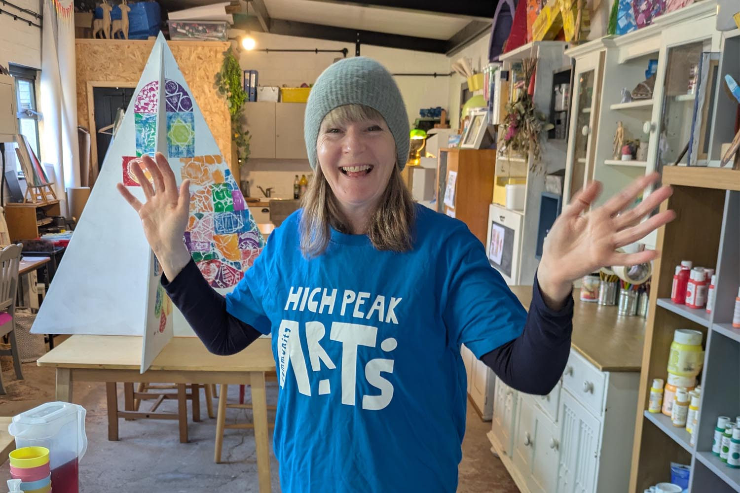 A woman smiling and waving, wearing a blue logo "High Peak Community Arts" t-shirt. In the background, a triangular-shaped Christmas tree with hand-printed designs in different colours.