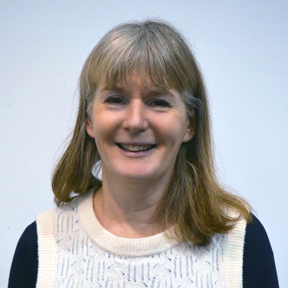 A woman with short hair and a fringe wearing a navy blue top with a cable knit vest over it.