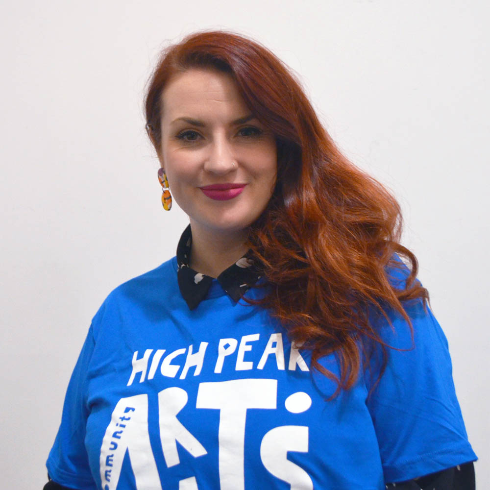 A woman with red hair, pink lipstick wearing a blue High Peak Community Arts logo t-shirt.