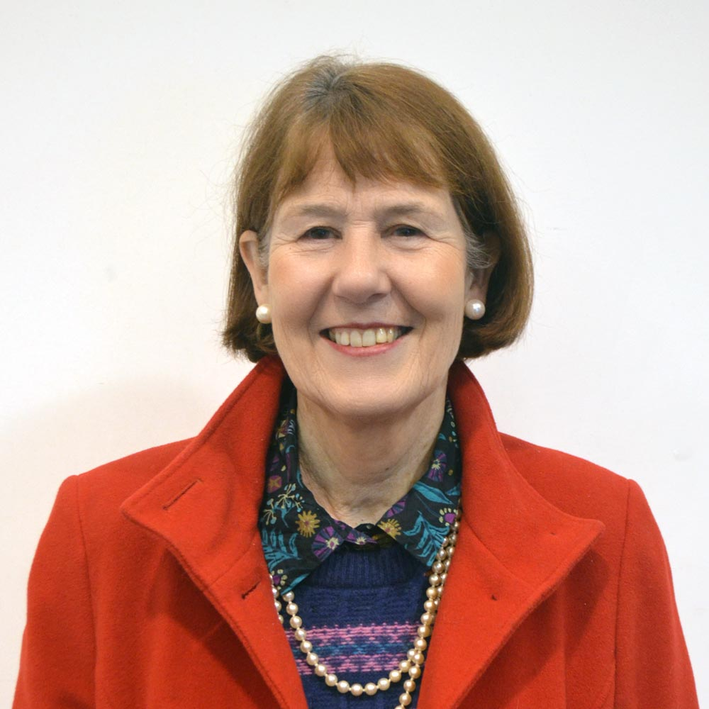 A woman with short hair and pearl earrings is smiling. She is wearing a red coat and a pearl bead necklace.
