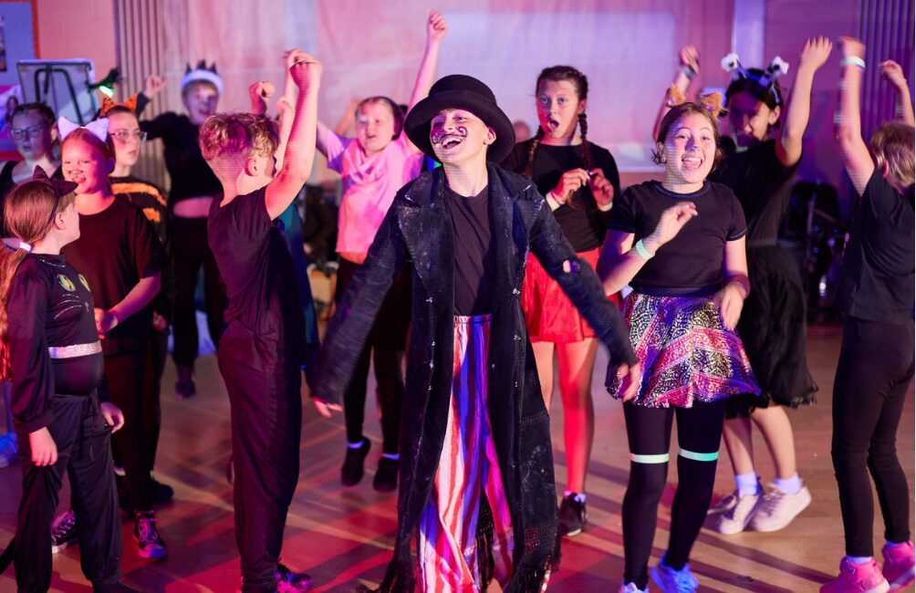 Young people dancing and smiling in different costumes such as: a ring master looking character with a drawn-on moustache, a glittery skirt, bunny ears, cat ears and a crown.