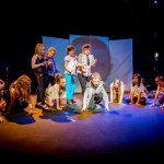 A blue‑lit stage with several young performers. Some are crouched and others are standing, holding their hands in claw‑shaped positions as part of the performance.
