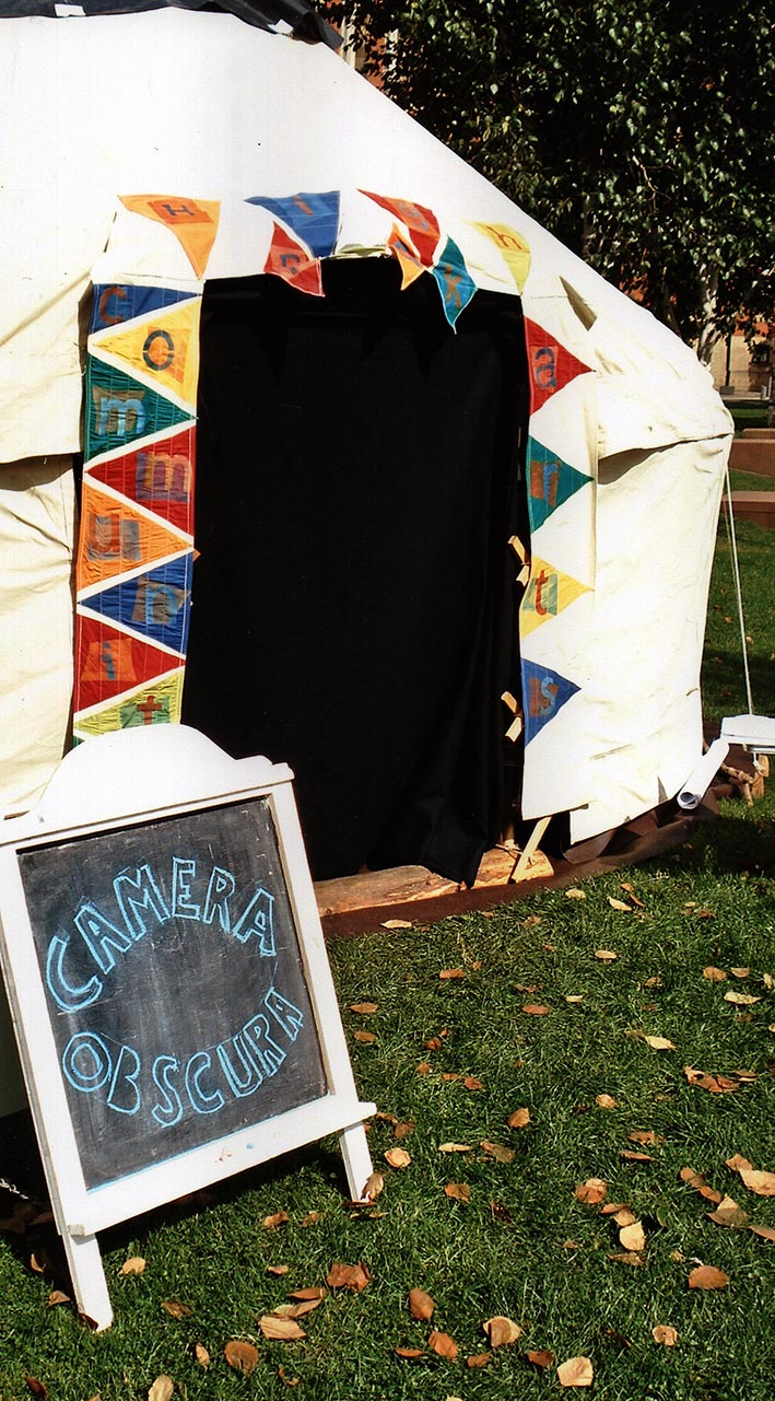A white Yurt on grass with a colourful bunting around the edge of the entrance. To the left of the entrance is a chalkboard with bubble writing saying "Camera Obscura".