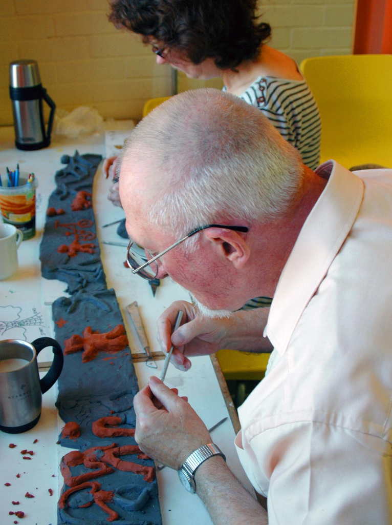 A male wearing a shirt and a lady in a black and white striped top in the background, working on a clay piece of art that has a blue base and orange people.
