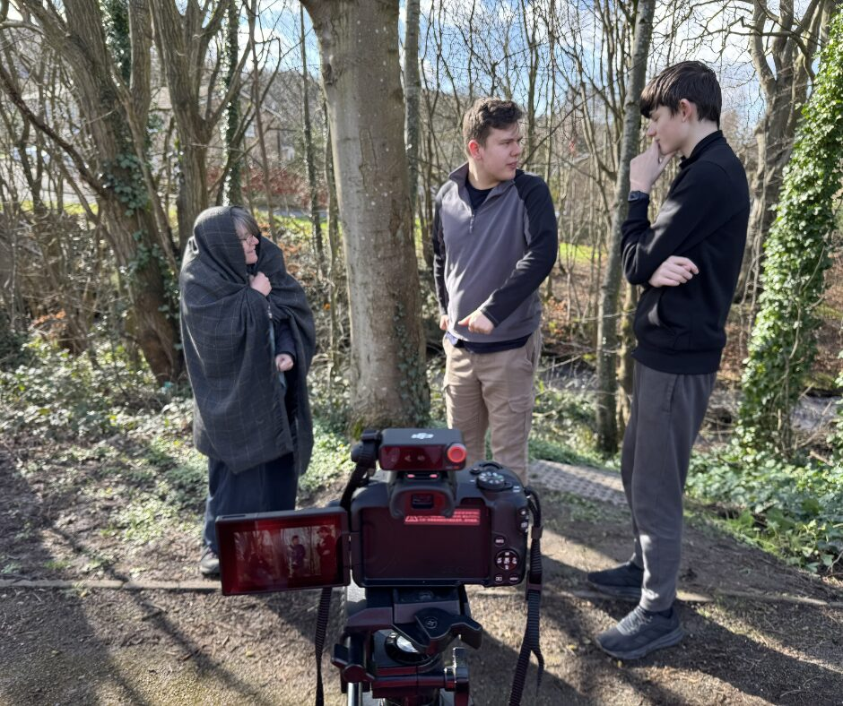 An outdoor shot filmed by a river, with 2 young men standing and a woman playing the part of an old lady talking together.