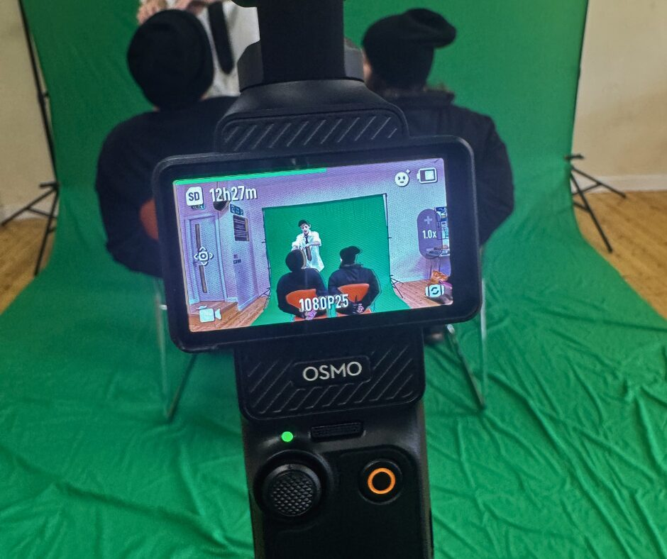 A photo of a camera shot showing a scene filmed on a green screen; in the scene, a person wearing a shirt and tie stands over 2 individuals in all-black clothing tied to chairs.