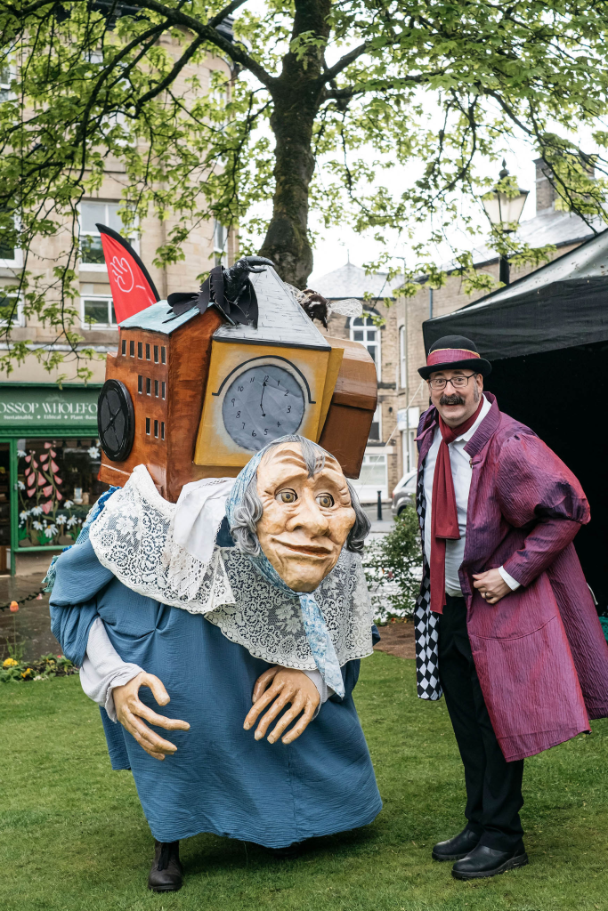 a puppet of an old lady with a mill on her back and a performer with a purple Victorian style outfit