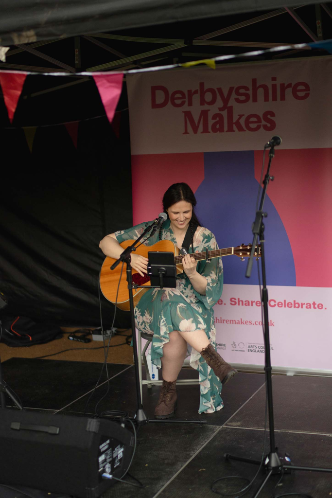 A female playing guitar at a festival with a green dress and brown boots