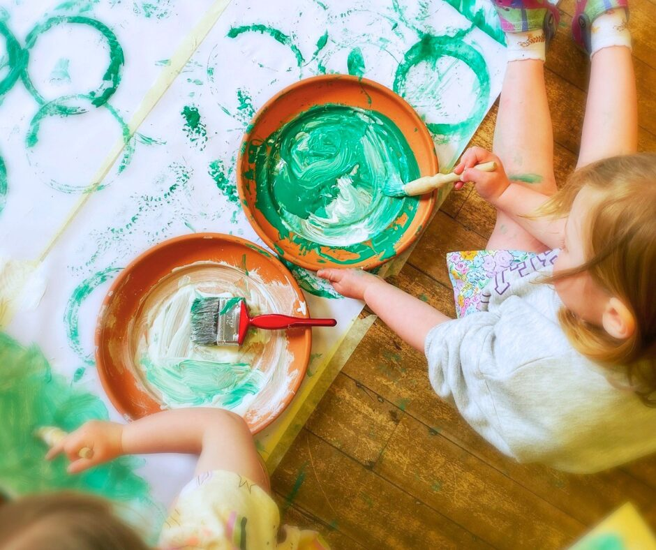 2 young children sat on the floor painting with green paint on a large sheet of paper.