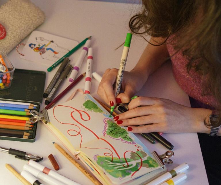 A person using vibrant-coloured pens and pencils to create artistic designs featuring trees and bushes in a sketch book.