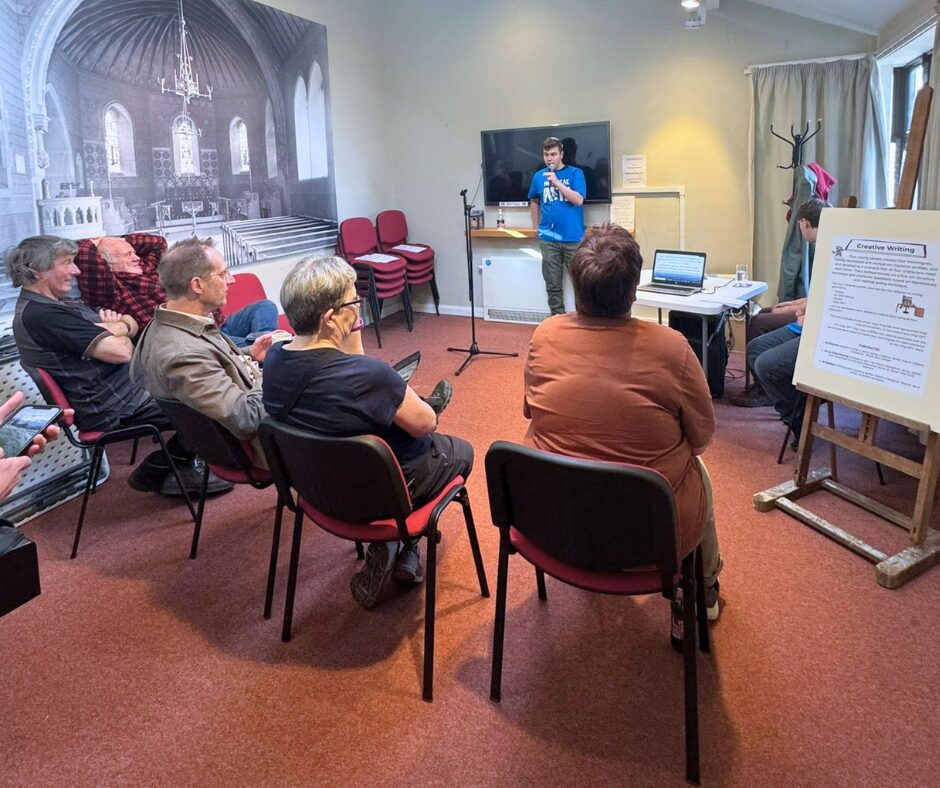 An audience of people sat watching a young man with a microphone speaking, wearing a blue High Peak Community Arts t-shirt, reading creative writing.