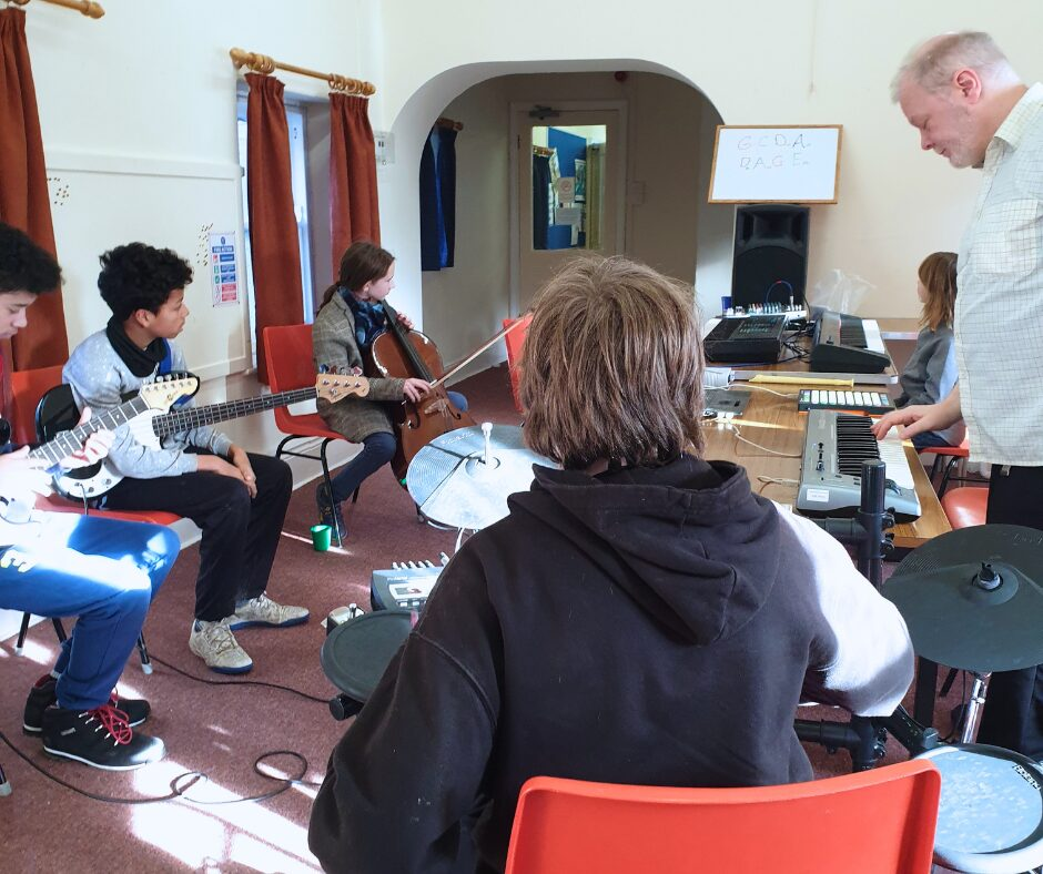 Young people playing various instruments, including guitars, a cello, an electronic drum kit and multiple keyboards. They are looking towards a whiteboard with the notes on.