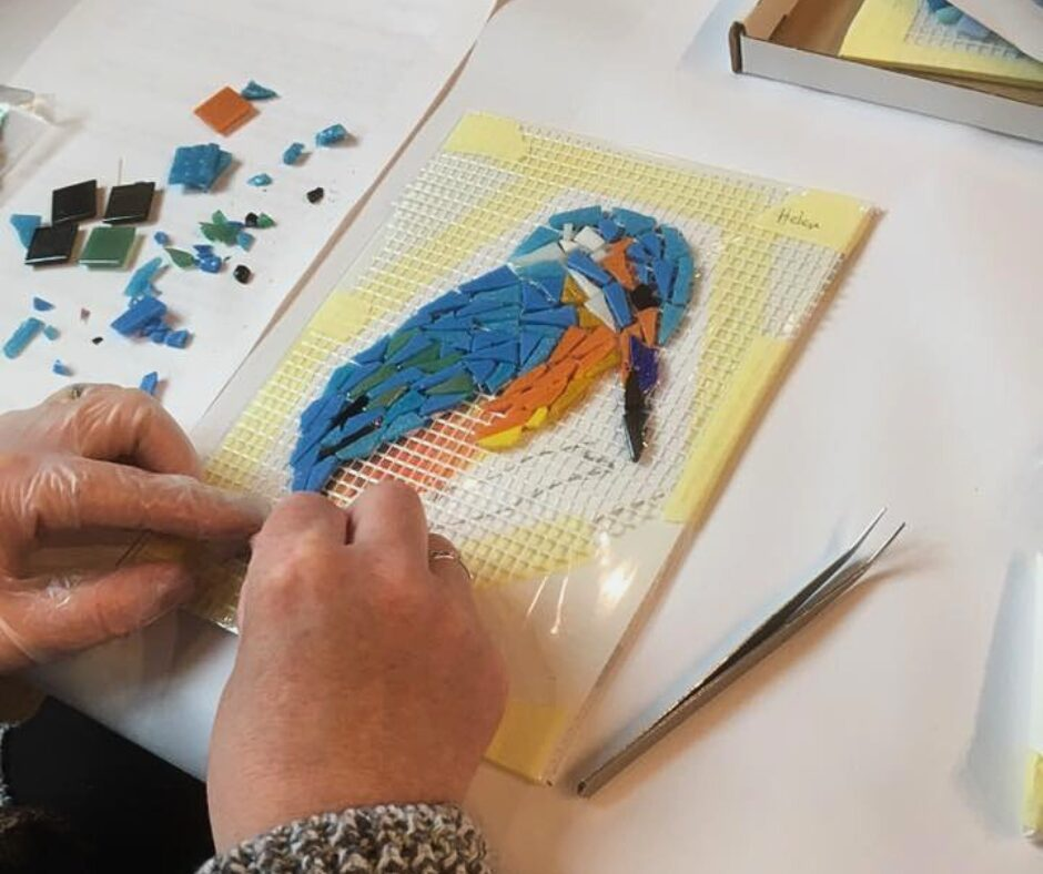 2 hands creating a classic bluebird with a bright orange belly mosaic made of cut pieces of glass.