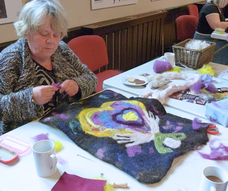 A woman holding purple wool by a large needle-felt project that features hands holding a heart.
