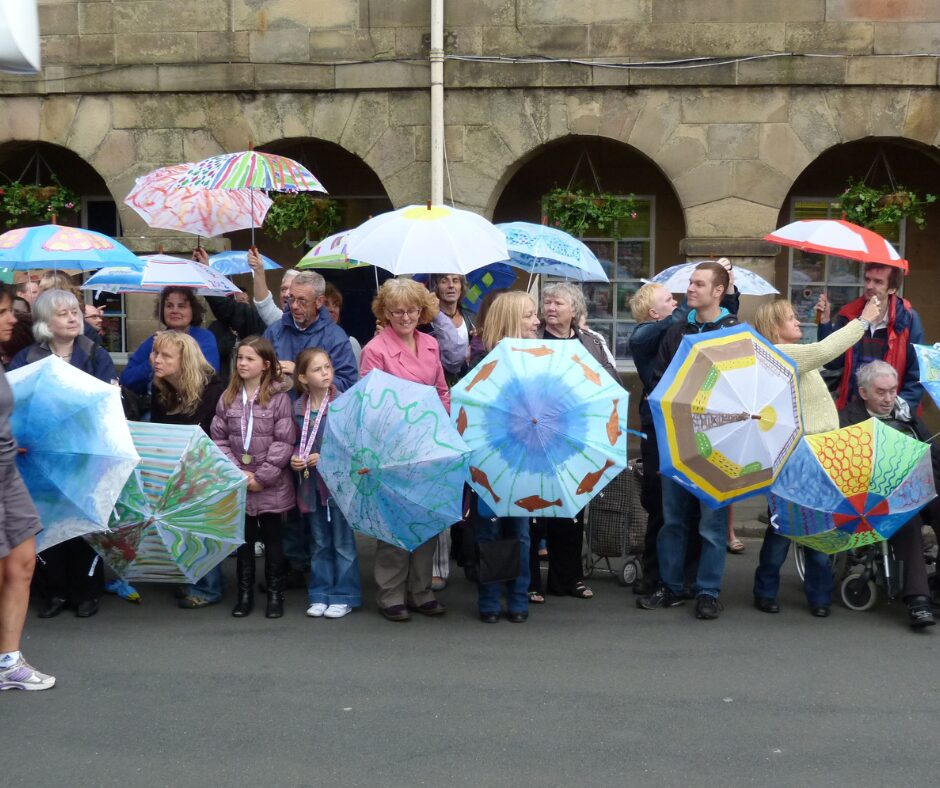 A group of people of all ages holding colourfully decorated umbrellas that feature fish, a lighthouse and other patterns made up of different shapes.