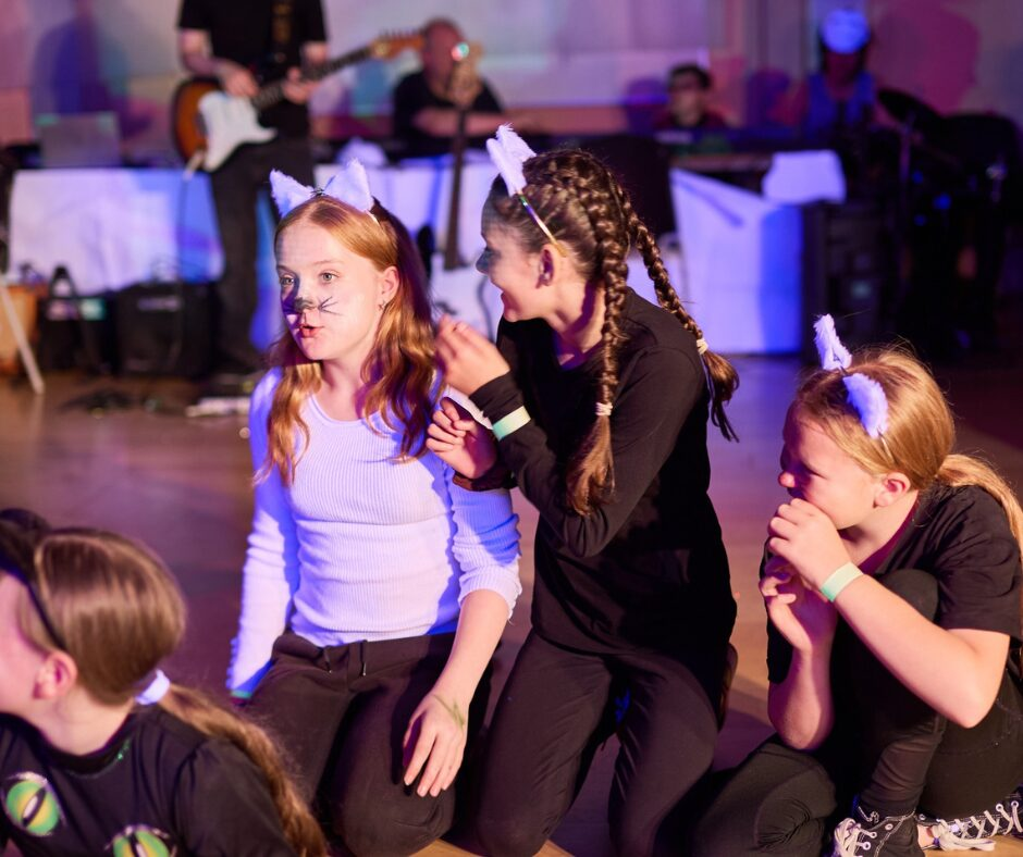 A group of young people kneeling, wearing cat ears on stage; in the background a band can be seen.
