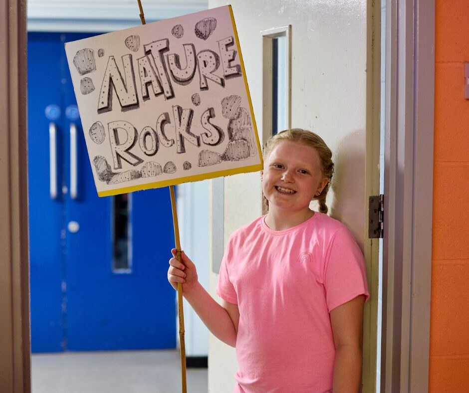 A young girl wearing a bright pink t-shirt is holding a sign on a stick that says "Nature Rocks" and rock drawings surrounding the text.